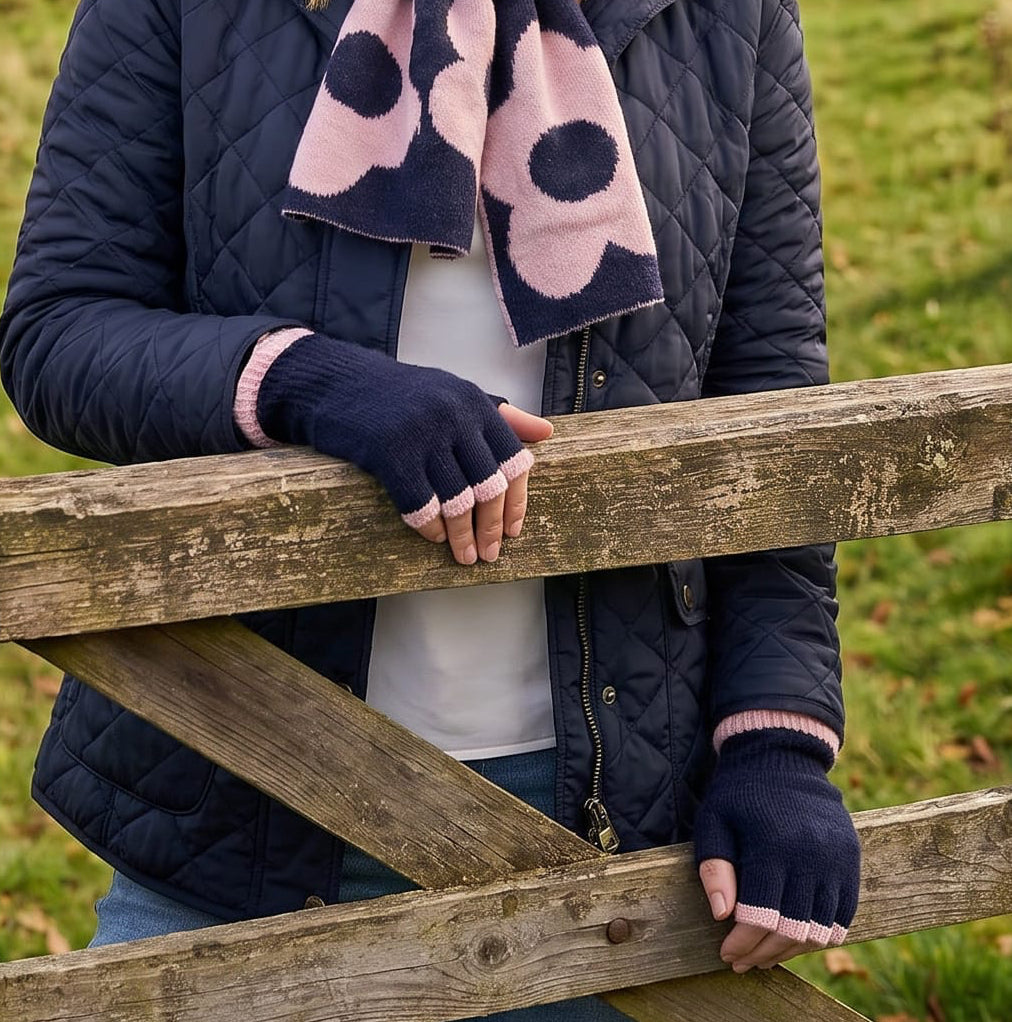 A woman wearing a navy blue beanie, navy quilted jacket, and a pink and navy patterned scarf leans on a wooden fence in a rural landscape with stone walls and green hills under a cloudy sky.