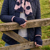A woman wearing a navy blue beanie, navy quilted jacket, and a pink and navy patterned scarf leans on a wooden fence in a rural landscape with stone walls and green hills under a cloudy sky.